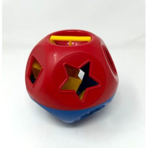 Puede incluir: Un juguete clasificador de formas rojo, azul y amarillo. El juguete es un cubo redondeado con varios recortes geométricos, incluyendo una estrella, un círculo y un triángulo. Una barra amarilla está en la parte superior. El juguete está diseñado para el desarrollo de la primera infancia.