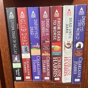 May include: A shelf of nine paperback books with red, black, and blue covers. The books are all by Charlaine Harris and are part of the Sookie Stackhouse series. The titles of the books are "Living Dead in Dallas", "Glib Dead", "Definitely Dead", "Dead as a Doornail", "To Worse", "From Dead", "Dead Until Dark", "Dead to the World".