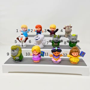 May include: A collection of twelve small plastic character figurines arranged on a three-tiered white display stand. Each figure is numbered from 1 to 12, featuring various animated characters with distinct outfits, expressions, and accessories.