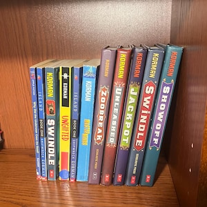 May include: A bookshelf with a collection of 11 paperback books by author Gordon Korman. The books are arranged in a row from left to right. The titles of the books are "Island", "Ungifted", "Masterminds", "Zoobreak", "Unleashed", "Jackpot", "Swindle", "Showoff", "Why Did the Underwear Cross the Road?", "Island", and "Swindle".