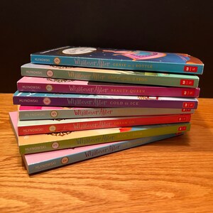 May include: A pile of children's books from the "Whatever After" series. The books have vibrant covers in shades of blue, pink, purple, and red. Titles include "Genie in a Bottle" and "Beauty Queen." The books are stacked on a wooden surface.