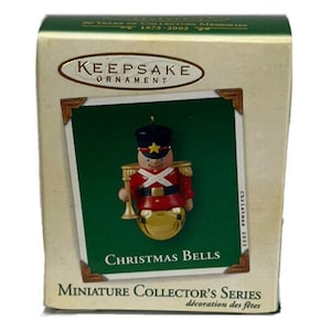 May include: A red and gold Christmas ornament featuring a toy soldier holding a bell. The ornament is part of the Hallmark Keepsake Ornament series and is labeled "Christmas Bells".