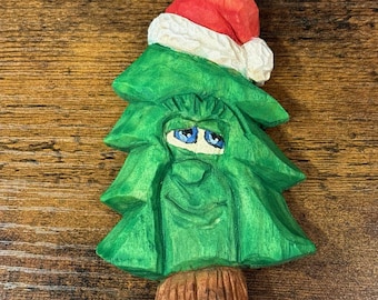 Puede incluir: Adorno de árbol de Navidad de madera tallada. El árbol verde tiene una cara sonriente con ojos azules y lleva un gorro de Papá Noel rojo con ribete blanco. La base del árbol es marrón. El adorno está diseñado para colgar.
