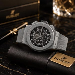Puede incluir: Un reloj Hublot gris con esfera negra y correa de caucho gris. El reloj se exhibe sobre un estuche de cuero negro con el logotipo dorado de Hublot. El reloj tiene un diseño complejo y detallado.