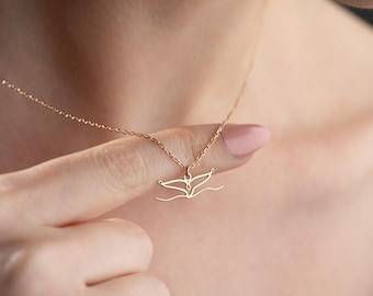 May include: A gold necklace with a whale tail pendant. The pendant is a minimalist design, showing the whale tail above a wavy line. The necklace is held by a finger, highlighting the pendant's fine details. The chain is delicate.