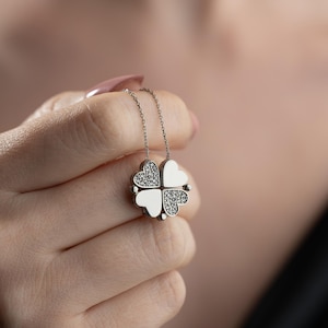 Peut inclure: Un collier en argent avec un pendentif en forme de trèfle à quatre feuilles. Chaque feuille en forme de cœur présente un design différent, avec deux feuilles ornées de petites pierres scintillantes et les deux autres de couleur blanche unie. Le collier est tenu dans une main.