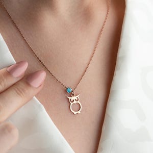 Puede incluir: Un collar de oro rosa con una cadena delicada y un colgante de búho. El búho está recortado y tiene una pequeña piedra preciosa azul en el centro.
