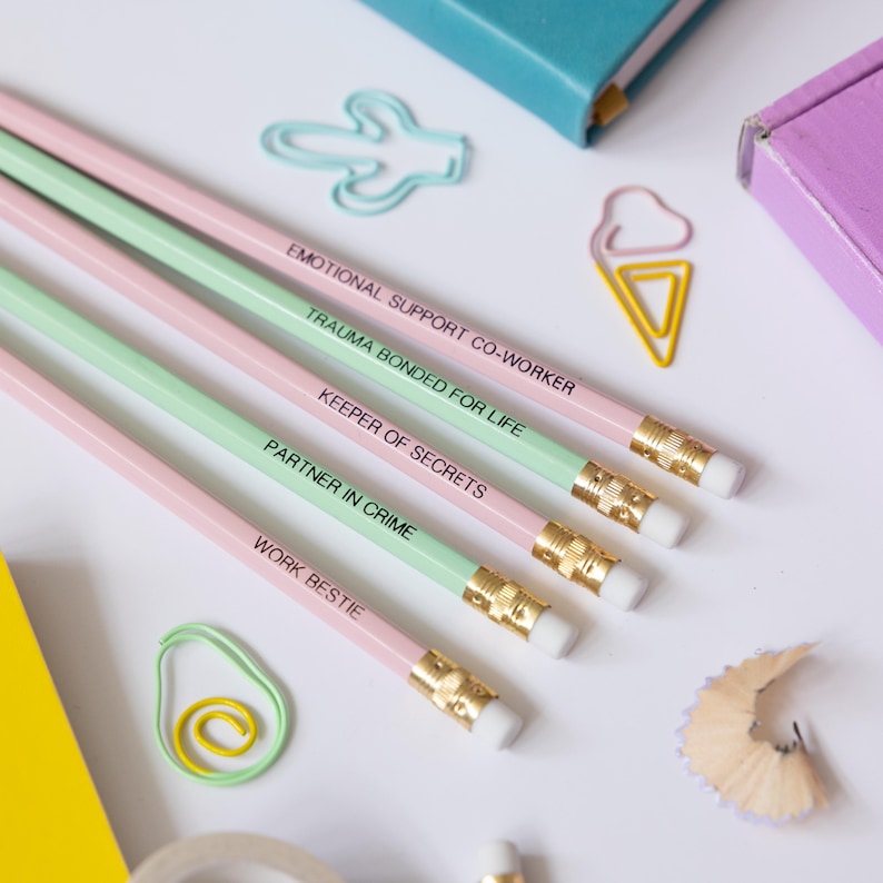 May include: Set of five pink and green pencils with funny phrases printed on them. The phrases are "Emotional Support Co-Worker", "Trauma Bonded For Life", "Keeper of Secrets", "Partner in Crime", and "Work Bestie".