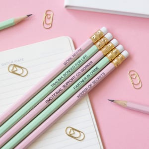 May include: Five pencils with different colored wood and black text on them. The text on the pencils reads "Work bestie", "Trauma bonded for life", "Emotional support co-worker", "Partner in crime", and "Keeper of secrets".