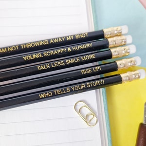 May include: A collection of black pencils with gold text, each inscribed with an inspirational phrase. The quotes include "I AM NOT THROWING AWAY MY SHOT," "YOUNG, SCRAPPY & HUNGRY," and "TALK LESS, SMILE MORE." The pencils have gold ferrules and white erasers.