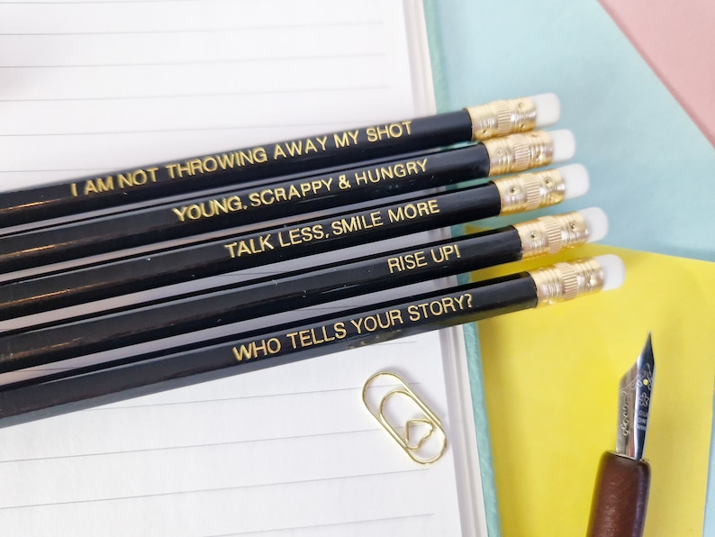 May include: A collection of black pencils with gold text, each inscribed with an inspirational phrase. The quotes include "I AM NOT THROWING AWAY MY SHOT," "YOUNG, SCRAPPY & HUNGRY," and "TALK LESS, SMILE MORE." The pencils have gold ferrules and white erasers.