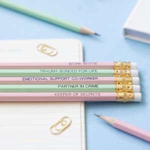 May include: Five pencils with pink and green painted wood and gold accents. The pencils have different phrases printed on them, including "Work Bestie", "Trauma Bonded for Life", "Emotional Support Co-Worker", "Partner in Crime", and "Keeper of Secrets".