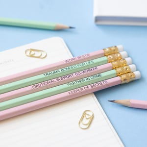 May include: Set of five pink and green pencils with gold accents. The pencils have different phrases printed on them, including "Work Bestie", "Trauma Bonded for Life", "Emotional Support Co-Worker", "Partner in Crime", and "Keeper of Secrets".