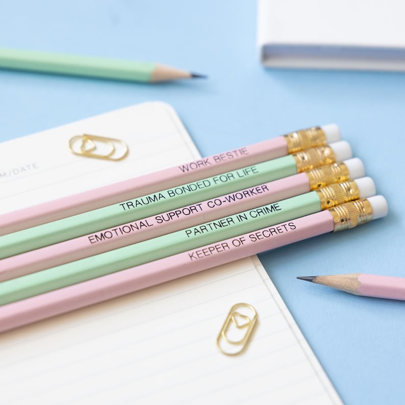 May include: Set of five pink and green pencils with gold accents. The pencils have different phrases printed on them, including "Work Bestie", "Trauma Bonded for Life", "Emotional Support Co-Worker", "Partner in Crime", and "Keeper of Secrets".