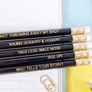 May include: A set of black pencils with gold lettering, each displaying an inspirational quote. The quotes include "I AM NOT THROWING AWAY MY SHOT," "YOUNG, SCRAPPY & HUNGRY," and "TALK LESS, SMILE MORE." The pencils have white erasers and gold-coloured metal bands.