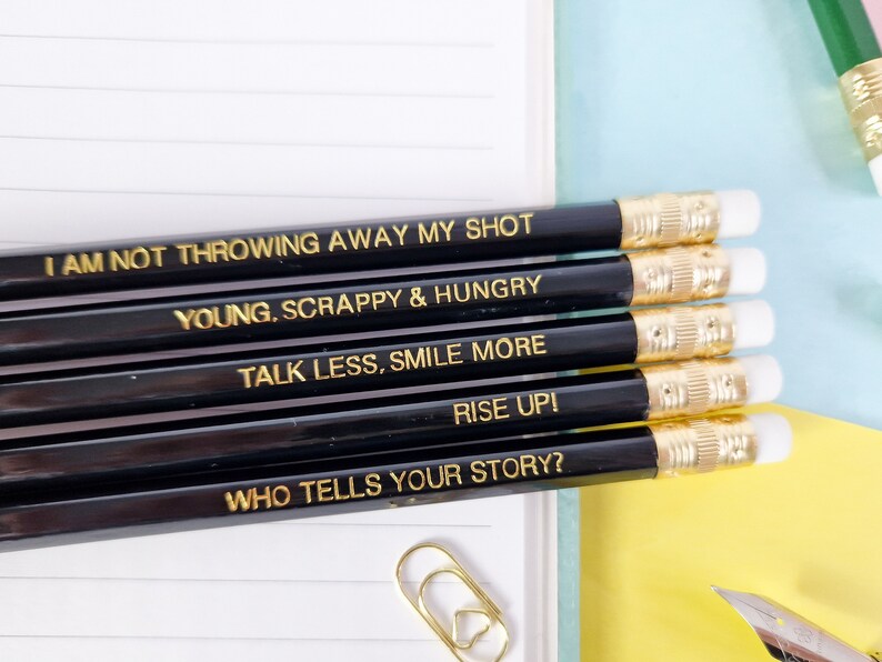 May include: A set of black pencils with gold lettering, each displaying an inspirational quote. The quotes include "I AM NOT THROWING AWAY MY SHOT," "YOUNG, SCRAPPY & HUNGRY," and "TALK LESS, SMILE MORE." The pencils have white erasers and gold-coloured metal bands.