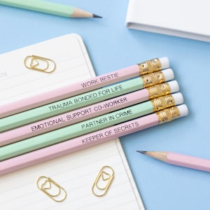 May include: Five pink and green pencils with different friendship-themed phrases written on them. The phrases are "Work Bestie", "Trauma Bonded For Life", "Emotional Support Co-Worker", "Partner In Crime", and "Keeper Of Secrets".