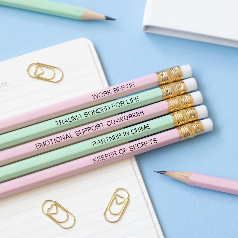 May include: Five pink and green pencils with different friendship-themed phrases written on them. The phrases are "Work Bestie", "Trauma Bonded For Life", "Emotional Support Co-Worker", "Partner In Crime", and "Keeper Of Secrets".