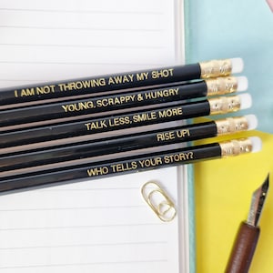 May include: A set of black pencils with gold lettering, each displaying an inspirational quote. The quotes include "I AM NOT THROWING AWAY MY SHOT," "YOUNG, SCRAPPY & HUNGRY," and "TALK LESS, SMILE MORE." The pencils have white erasers and gold metal bands.