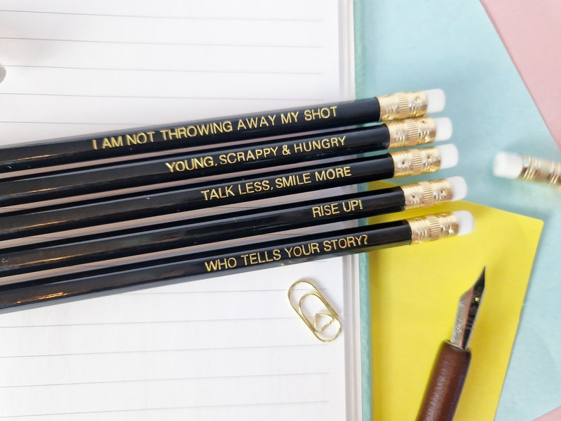 May include: A set of black pencils with gold lettering, each displaying an inspirational quote. The quotes include "I AM NOT THROWING AWAY MY SHOT," "YOUNG, SCRAPPY & HUNGRY," and "TALK LESS, SMILE MORE." The pencils have white erasers and gold metal bands.