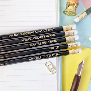 May include: A selection of black pencils with gold lettering, each inscribed with an inspirational quote. The quotes include "I AM NOT THROWING AWAY MY SHOT," "YOUNG, SCRAPPY & HUNGRY," and "TALK LESS, SMILE MORE." The pencils are placed on a lined notebook.