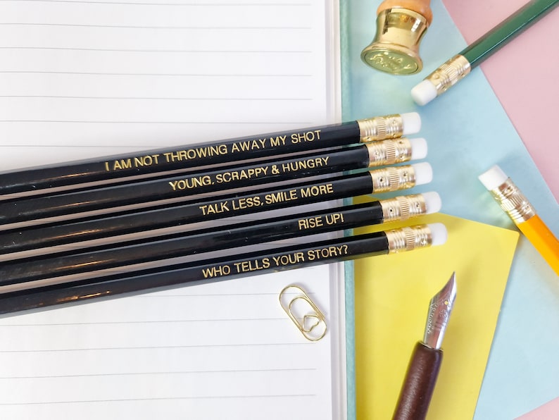 May include: A selection of black pencils with gold lettering, each inscribed with an inspirational quote. The quotes include "I AM NOT THROWING AWAY MY SHOT," "YOUNG, SCRAPPY & HUNGRY," and "TALK LESS, SMILE MORE." The pencils are placed on a lined notebook.