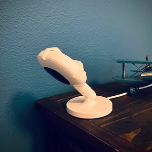 May include: A white baby monitor with a round camera lens and a white base. The monitor is on a dark brown wooden surface.