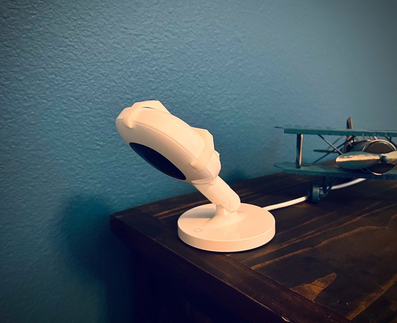 May include: A white baby monitor with a round camera lens and a white base. The monitor is on a dark brown wooden surface.