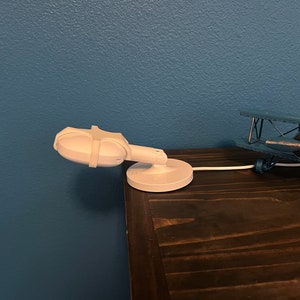 May include: A white baby monitor with a swivel base is mounted on a wooden table. The monitor is pointed towards the left side of the image. A blue toy airplane is on the table to the right of the monitor.