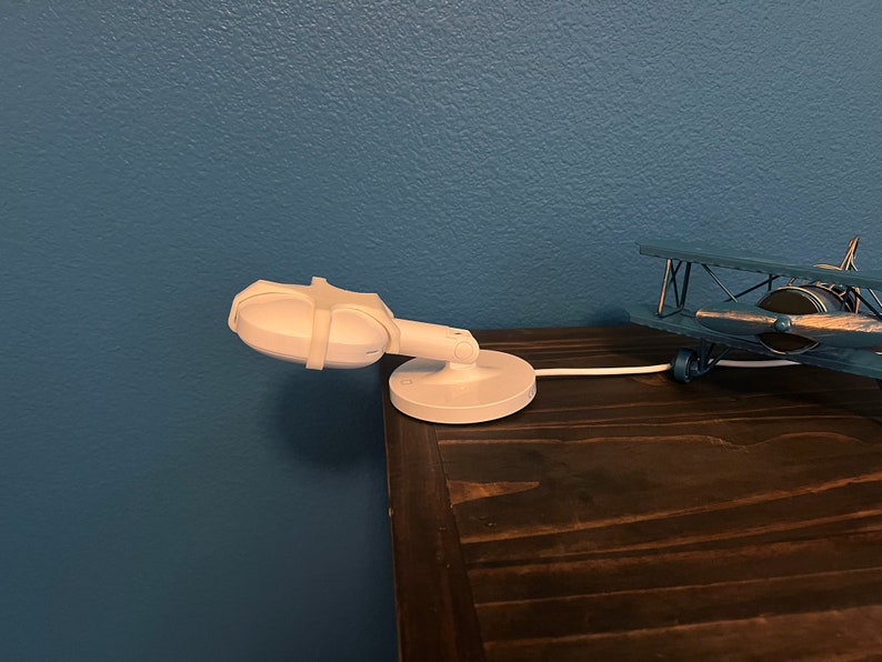 May include: A white baby monitor with a swivel base is mounted on a wooden table. The monitor is pointed towards the left side of the image. A blue toy airplane is on the table to the right of the monitor.