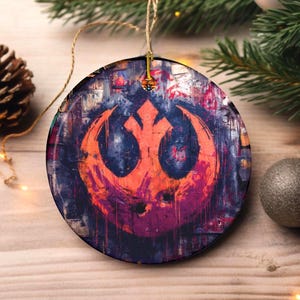 May include: Round Christmas ornament with a distressed, abstract design featuring an orange emblem on a dark blue and purple background. The ornament is hanging by a natural twine string. Festive holiday decor.