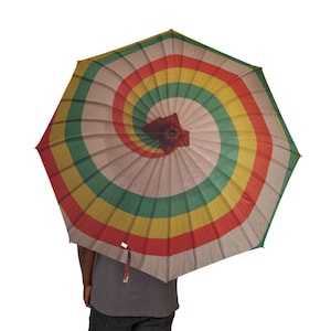 May include: A colorful umbrella with a spiral design. The umbrella features alternating bands of red, yellow, green, and tan. The umbrella is open, and the handle is not visible. The umbrella is held by a person wearing a gray shirt and dark pants.