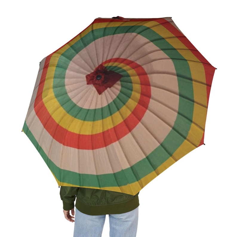 May include: A colorful umbrella with a spiral design. The umbrella features alternating bands of red, yellow, green, and beige. The umbrella is open, and the handle is not visible. The umbrella is held by a person wearing a green jacket and blue jeans.