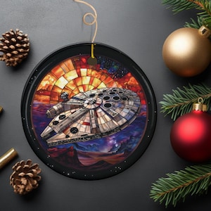 May include: A round, black, framed ornament with a stained glass design of the Millennium Falcon spaceship flying through a colorful nebula. The spaceship is white with gray details and has a circular window pattern. The background is a vibrant mix of orange, red, purple, and blue.