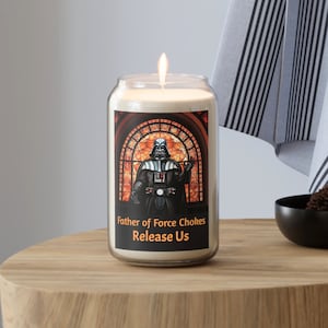 May include: A white candle in a glass jar with a black and white image of Darth Vader in a stained glass window. The text on the jar reads "Father of Force Chokes Release Us."