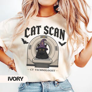 May include: An ivory t-shirt featuring a graphic of a black cat wearing a purple wizard hat with stars and a moon, sitting inside a CT scanner. The text reads CAT SCAN above the scanner and CT TECHNOLOGIST below, with two small black bats on sides.