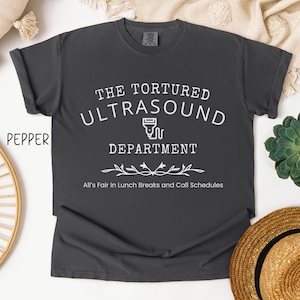 May include: A dark gray t-shirt with white text that reads "The Tortured Ultrasound Department" with a stylized ultrasound machine graphic and a floral design below. The text below the graphic reads "All's Fair in Lunch Breaks and Call Schedules."