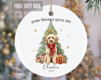 May include: White ceramic Christmas ornament with a watercolor illustration of a Doodle dog wearing a red scarf in front of a Christmas tree. Text reads "Some Doodle Loves You", "Charlie", and "2025". Gold stars and wrapped gifts are also featured.