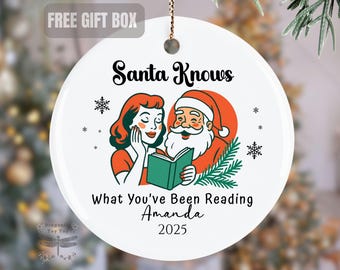 May include: White ceramic Christmas ornament with a retro illustration of Santa Claus and a woman reading a book. The text reads "Santa Knows What You've Been Reading Amanda 2025". Includes snowflakes and a gift box.