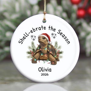 May include: A round white ceramic ornament featuring a brown tortoise wearing a red Santa hat, wrapped in colorful string lights, with pine branches and berries. The text reads Shell-ebrate the Season, Olivia, and 2026 in black font.