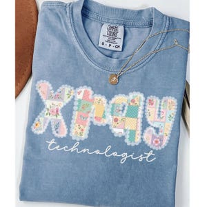 May include: A light blue Comfort Colors t-shirt with the word "XRAY" in a patchwork design with floral and butterfly patterns. The word "technologist" is written below in a script font. A gold necklace with a pendant is draped on the shirt.