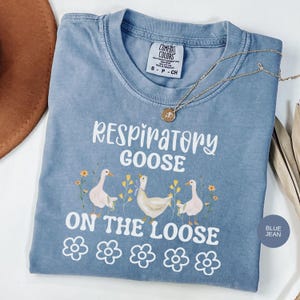 May include: A blue jean Comfort Colors t-shirt with the phrase "Respiratory Goose on the Loose" in white. The shirt features illustrations of geese and flowers. A gold necklace with a pendant is draped across the shirt.