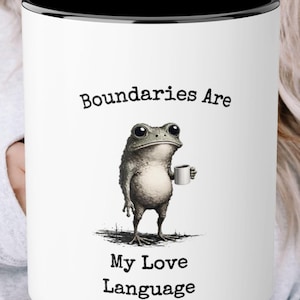 May include: A white ceramic mug with a black interior rim featuring a black illustration of a standing frog holding a small white mug. The text "Boundaries Are My Love Language" is printed in black typewriter-style font above and below the frog.
