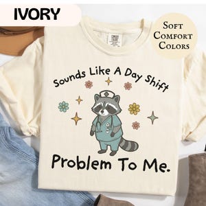 May include: Ivory-colored t-shirt with the text "Sounds Like A Day Shift Problem To Me." features a cartoon raccoon wearing a nurse's uniform. The shirt also has floral and star graphics. The words "Soft Comfort Colors" are also on the shirt.