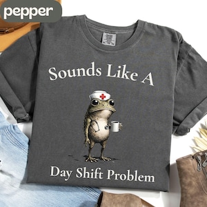 May include: Dark gray t-shirt with the text "Sounds Like A Day Shift Problem." The shirt features a cartoon frog wearing a nurse's hat and holding a coffee cup. The shirt is a crew neck style.
