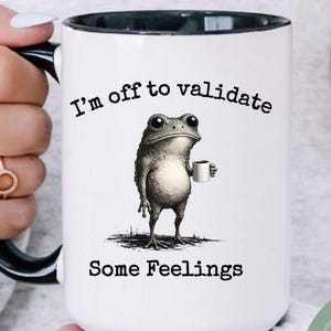 May include: White ceramic mug with a black interior and handle. The mug features a whimsical illustration of a frog holding a coffee cup, with the text "I'm off to validate Some Feelings."