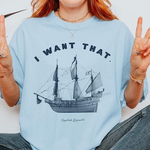 May include: A light blue t-shirt featuring the text "I WANT THAT." and a vintage ship illustration. The ship is labelled "Napoleon Dynamite".