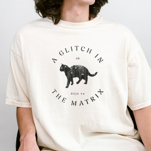 May include: A cream colored t-shirt with a black cat graphic and the text "A Glitch in the Matrix" with the words "oh" and "Déjà vu" in smaller font.
