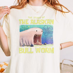 May include: Cream-colored t-shirt featuring a cartoon illustration of a pink worm with the text "THE STORY OF THE ALASKAN BULL WORM" in yellow. The graphic is centered on the front of the shirt.