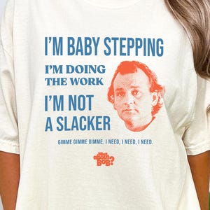 May include: Ivory-colored t-shirt with the text "I'M BABY STEPPING, I'M DOING THE WORK, I'M NOT A SLACKER" in blue. The shirt features a portrait of a man with orange hair and the phrase "GIMME GIMME GIMME. I NEED, I NEED, I NEED."